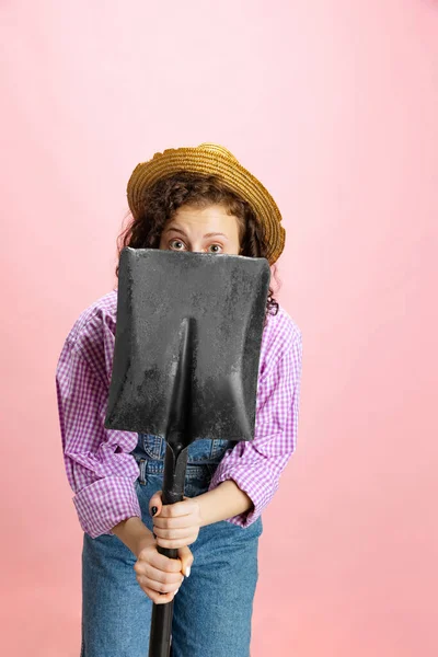 Komiczny portret młodej wystraszonej dziewczyny, ogrodnika w mundurze roboczym i słomiany kapelusz chowający się za łopatą odizolowany na różowym tle. Pojęcie pracy, emocji, agronomii, zabawy — Zdjęcie stockowe