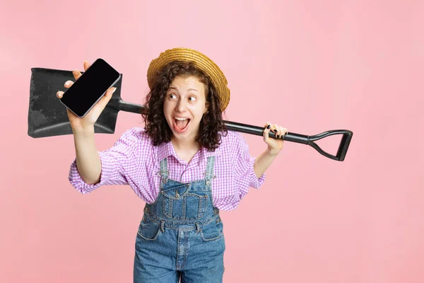 Studio girato di giovane ragazza felice, giardiniere donna in uniforme da lavoro e cappello utilizzando telefono isolato su sfondo rosa. Concetto di lavoro, emozioni, agronomia, eco — Foto Stock