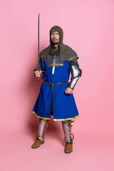 Ganztägiges Porträt eines ernst aussehenden Mannes, mittelalterlichen Kriegers oder Ritters in Schutzhülle mit Schwert isoliert über rosa Studiohintergrund — Stockfoto