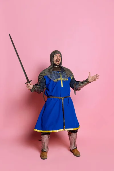Pełnowymiarowy portret człowieka odważnego średniowiecznego wojownika lub rycerza w łańcuszkowej zbroi z mieczem krzyczącym na różowym tle studia — Zdjęcie stockowe
