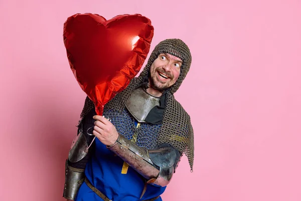 Halblanges Porträt eines glücklichen und aufgeregten Mannes, mittelalterlicher Krieger in Schutzhülle mit rotem Ballon in Herzform, isoliert über rosa Hintergrund — Stockfoto