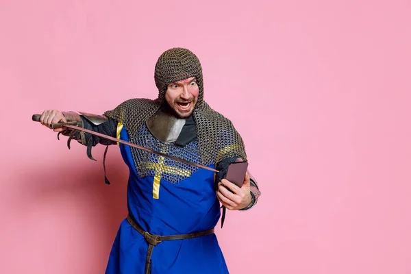 Retrato de meio-comprimento do homem, guerreiro medieval ou cavaleiro de armadura apontando espada na tela do telefone isolado sobre fundo de estúdio rosa — Fotografia de Stock