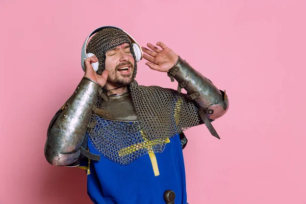 Retrato de meio-comprimento do homem, guerreiro medieval ou cavaleiro ouvindo música em fones de ouvido com expressão deliciosa isolado sobre fundo rosa — Fotografia de Stock