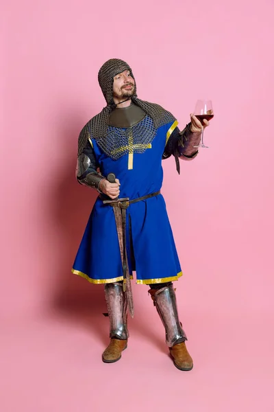 Full längd porträtt av människan med härlig ansiktsuttryck i amor med svärd, hålla glas av vinstockar isolerad över rosa bakgrund — Stockfoto