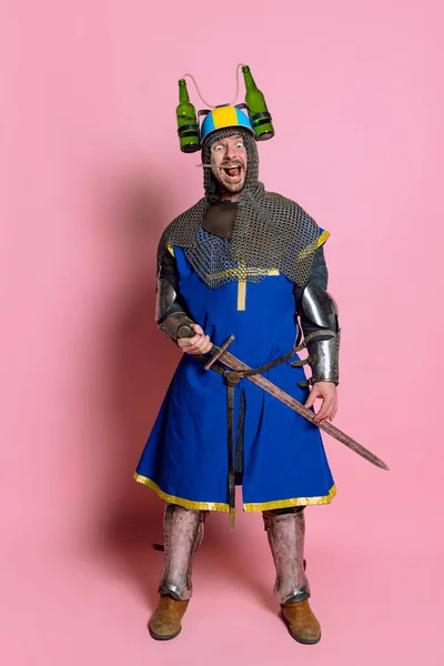 Retrato completo do homem, guerreiro medieval de armadura, com espada usando capacete de cerveja e fazendo rostos engraçados isolados sobre fundo rosa — Fotografia de Stock