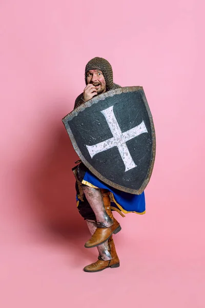 Pełnowymiarowy portret przerażającego człowieka, średniowiecznego wojownika lub rycerza w zbroi ukrywającego się za tarczą odizolowaną na różowym tle studia — Zdjęcie stockowe