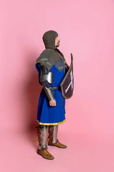 Πλάγια άποψη πλήρους μήκους πορτρέτο του γενναίου άνδρα, μεσαιωνικού πολεμιστή ή ιππότη σε προστατευτική πανοπλία με ασπίδα απομονωμένη πάνω από ροζ φόντο στούντιο — Φωτογραφία Αρχείου
