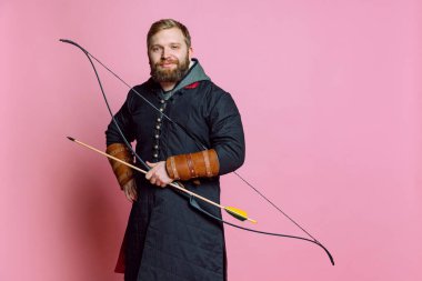 Orta çağ savaşçısı suretinde gülümseyen sakallı genç bir adamın stüdyo resmi. Pembe stüdyo arka planında izole edilmiş zırhlı okçuluk.