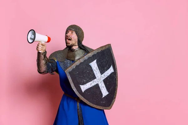 Retrato de homem, guerreiro medieval ou cavaleiro de armadura com escudo gritando em megafone isolado sobre fundo de estúdio rosa — Fotografia de Stock