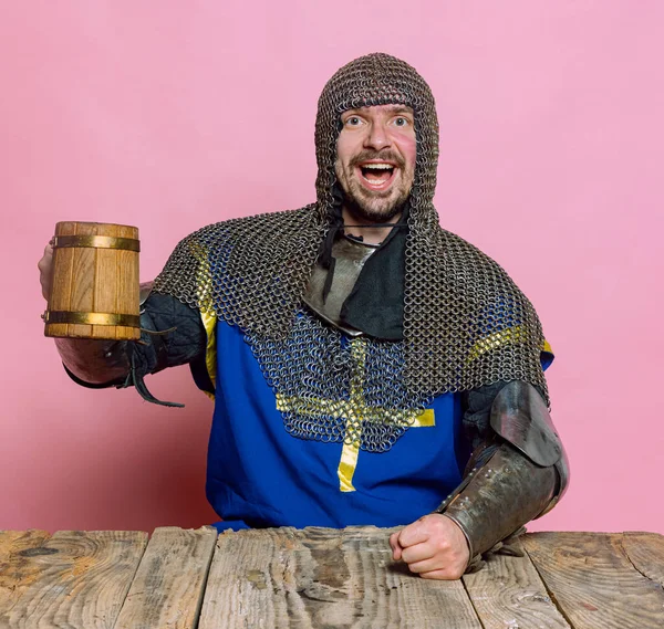 Portret podnieconego mężczyzny, średniowieczny wojownik, rycerz w specjalnej ochronie wznoszący się drewniany kubek na różowym tle studia — Zdjęcie stockowe