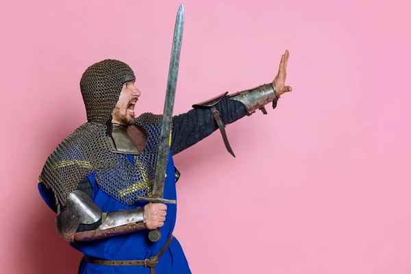Portret odważnego człowieka, średniowiecznego wojownika lub rycerza w zbroi z podnoszącą się ręką z mieczem pokazującą gest stop na różowym tle pracowni — Zdjęcie stockowe