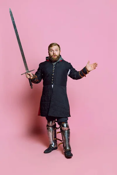 Ganztägiges Porträt eines jungen bärtigen Mannes, mittelalterlicher Krieger oder Bogenschütze im Schutz mit Schwert isoliert über rosa Hintergrund — Stockfoto
