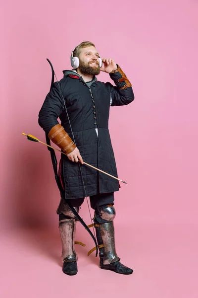 Портрет молодого бородатого человека в образе средневекового воина, лучника в цепной броне, слушающего музыку в наушниках, изолированных на розовом фоне — стоковое фото