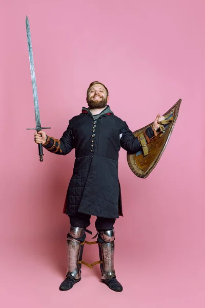 Ganztägiges Porträt eines jungen lächelnden Mannes, mittelalterlicher Krieger oder Bogenschütze in Rüstung mit Schwert und Schild isoliert über rosa Hintergrund — Stockfoto