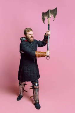Ortaçağ savaşçısı suretinde genç sakallı bir adamın atölye fotoğrafı pembe arka planda izole edilmiş baltalı zırhlı okçuluk. Dönem Karşılaştırması