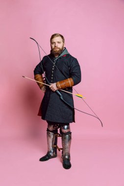 Orta çağ savaşçısı suretinde sakallı genç bir adamın stüdyo fotoğrafı. Pembe arka planda izole edilmiş eski moda bir kostüm giyen okçuluk..