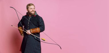 Orta çağ savaşçısı suretinde gülümseyen sakallı genç bir adamın stüdyo fotoğrafı. Pembe arka planda izole edilmiş eski moda kostümler giyen okçuluk. El ilanı