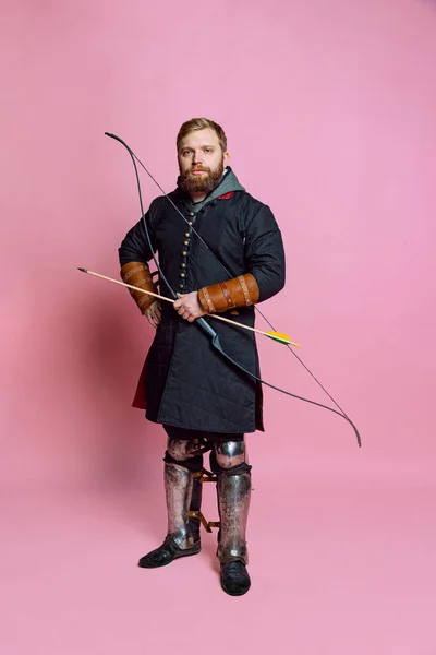 中世の戦士をイメージした若い髭を生やした男のスタジオショット、ピンクの背景に隔離されたヴィンテージ時代の衣装を身に着けているアーチェリー. — ストック写真