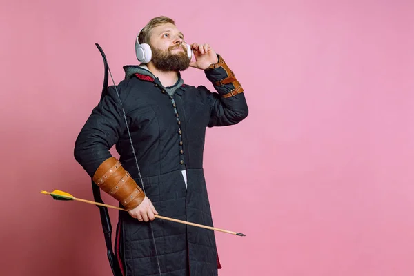Portré fiatal szakállas férfi képére középkori harcos, íjász lánc páncél hallgat zenét fülhallgató elszigetelt rózsaszín háttér — Stock Fotó