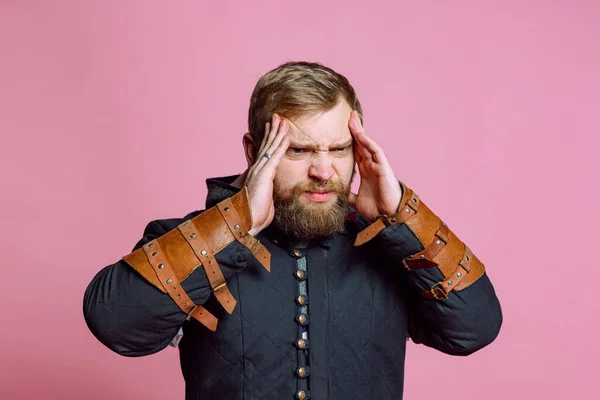 Retrato de joven barbudo en imagen de guerrero medieval, arquero sufriendo de dolor, tocando templos en dolor de cabeza aislado sobre fondo rosa — Foto de Stock