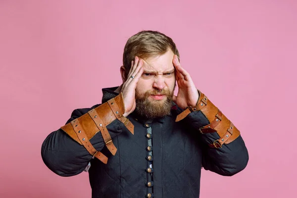 Porträt eines jungen bärtigen Mannes im Bild eines mittelalterlichen Kriegers, Bogenschütze mit Schmerzen, Berührung der Schläfen unter Kopfschmerzen isoliert vor rosa Hintergrund — Stockfoto