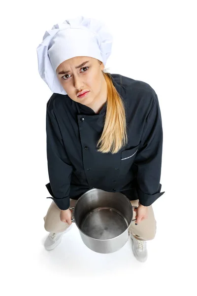 Jovencita triste, cocinera, cocinera de uniforme negro sosteniendo una cacerola grande aislada sobre fondo blanco. Cocina, profesión, negocio, comida, gusto, concepto de restaurante. —  Fotos de Stock