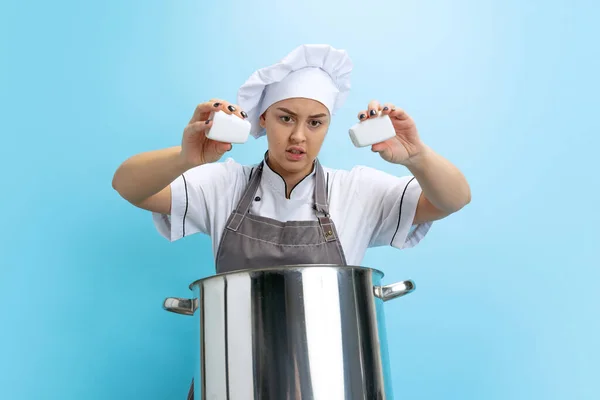 Ritratto di giovane donna caucasica, cuoca, cuoca in uniforme bianca con casseruola isolata su sfondo blu. Divertenti emozioni meme — Foto Stock