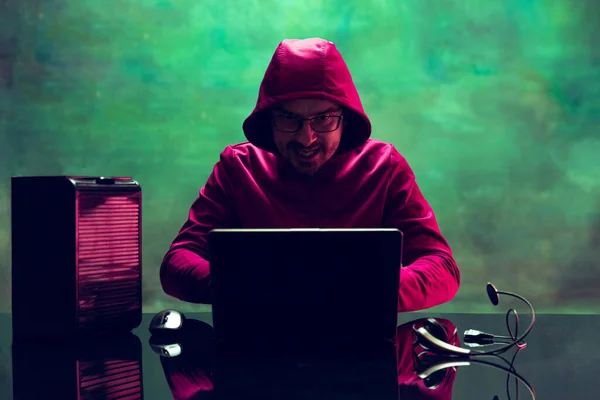 Portrait of man, computer geek, hacker working with laptop, breaking security system isolated over dark green background in neon — Stok Foto