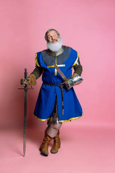 ピンクの背景に隔離された剣と装甲服のシニアグレーの髭の男、勇敢な中世の戦士や騎士の肖像画。時代、歴史、祭りの比較 — ストック写真