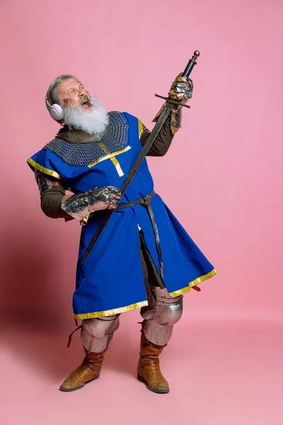 Retrato de homem barbudo cinza sênior, bravo guerreiro medieval ou cavaleiro em roupas blindadas com espada isolada em fundo rosa. Comparação de eras, história, festival — Fotografia de Stock