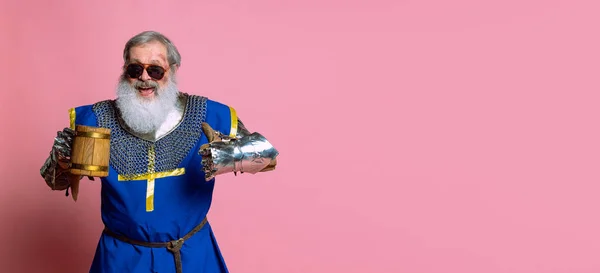 Retrato de homem barbudo cinza sênior, bravo guerreiro medieval ou cavaleiro em roupas blindadas com espada isolada em fundo rosa. Comparação de eras, história, festival — Fotografia de Stock