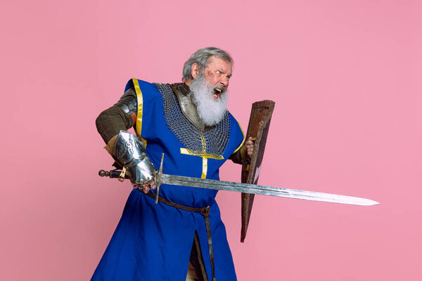Portrait of senior grey bearded man, brave medieval warrior or knight in armored clothes with sword isolated on pink background. Comparison of eras, history, festival
