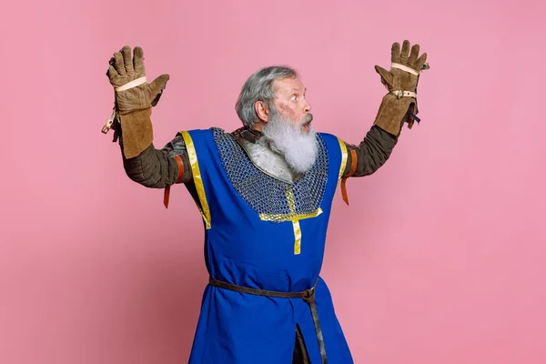 Portrait of senior grey bearded man, brave medieval warrior or knight in armored clothes with sword isolated on pink background. Comparison of eras, history, festival — Stock Photo, Image