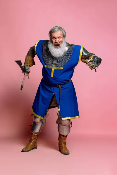Portret starszego siwego brodatego mężczyzny, dzielnego średniowiecznego wojownika lub rycerza w opancerzonych ubraniach z mieczem odizolowanym na różowym tle. Porównanie epok, historii, festiwalu — Zdjęcie stockowe