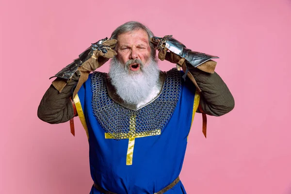 Porträt eines älteren Mannes mit grauem Bart, tapferer mittelalterlicher Krieger oder Ritter in gepanzerten Gewändern und Schwert isoliert auf rosa Hintergrund. Vergleich von Epochen, Geschichte, Festen — Stockfoto