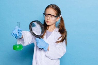 Küçük liseli bir kızın portresi, büyük beyaz cüppeli bir çocuk kimyager, kimyasal ekipman bilim adamı, labaratuarda sıvı, mavi arka planda izole edilmiş.
