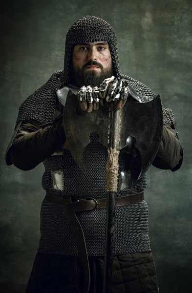Tradiční portrét brutálního seriózního muže, středověkého válečníka nebo rytíře se zraněnou tváří v kroužkové zbroji, držícího sekeru izolovanou nad tmavým pozadím. Porovnání období — Stock fotografie
