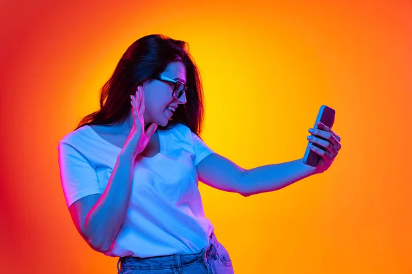 Young beautiful caucasian girl with long dark hair using phone isolated on orange background in neon light, filter. Concept of emotions, ads, sale — Stock Photo, Image