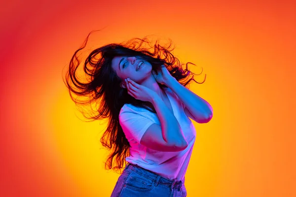Garota caucasiana bonita excitada com longos cabelos escuros dançando isolado em fundo laranja em luz de néon, filtro. Conceito de emoções, anúncios, venda — Fotografia de Stock