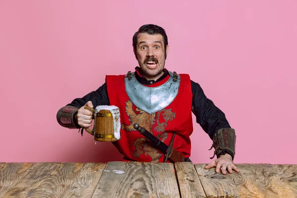 Porträt eines mittelalterlichen Kriegers oder Ritters mit schmutzigem, verwundetem Gesicht, der einen riesigen Bierkrug isoliert vor rosa Hintergrund hält. Vergleich von Epochen, Geschichte — Stockfoto