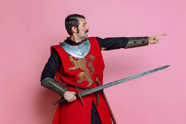 Komiczny portret średniowiecznego wojownika lub rycerza noszącego zbroję trzymającego duży miecz na różowym tle. Porównanie epok, historii — Zdjęcie stockowe