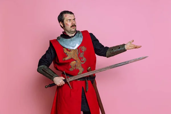 Retrato cômico de guerreiro medieval ou cavaleiro vestindo roupas de armadura segurando grande espada isolada sobre fundo rosa. Comparação de eras, história — Fotografia de Stock