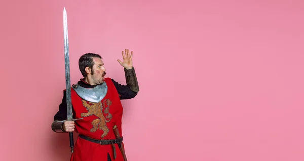 Portret zszokowanego średniowiecznego wojownika lub rycerza ubranego w zbroję trzymającego duży miecz na różowym tle. Porównanie epok, historii — Zdjęcie stockowe