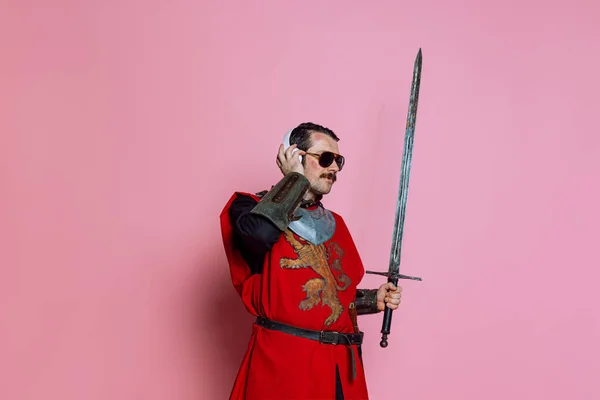 Studio shot di uomo, guerriero medievale o cavaliere che ascolta musica in cuffia con deliziosa espressione isolata su sfondo rosa — Foto Stock