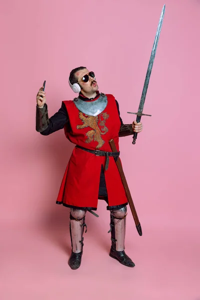 Studio shot di uomo, guerriero medievale o cavaliere che ascolta musica in cuffia con deliziosa espressione isolata su sfondo rosa — Foto Stock