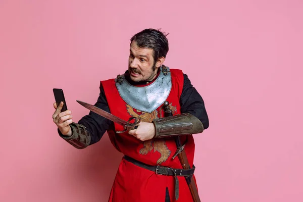 Κωμικό πορτραίτο μεσαιωνικού πολεμιστή ή ιππότη που φοράει πανοπλία χρησιμοποιώντας τηλέφωνο απομονωμένο σε ροζ φόντο. Σύγκριση εποχών, ιστορία — Φωτογραφία Αρχείου