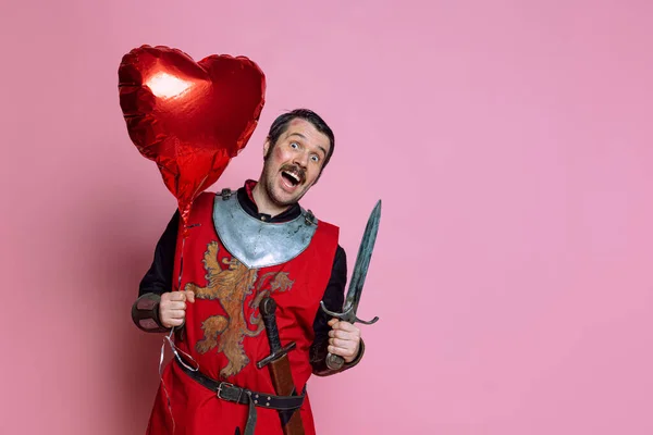 Połowa długości portret szczęśliwego i podekscytowanego człowieka, średniowieczny wojownik w powłoce ochronnej z czerwonym kształcie serca balon izolowany na różowym tle — Zdjęcie stockowe