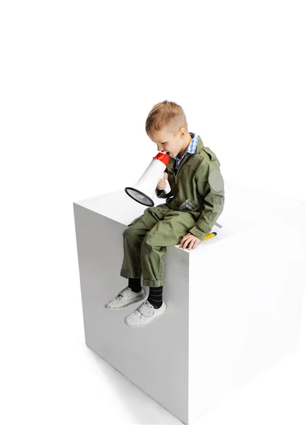 Ein kleiner Junge, niedliches Kind im Bild des Automechanikers oder Installateurs in grüner Latzhose mit Arbeitswerkzeug, das auf einer großen Box sitzt, isoliert über weißem Studiohintergrund. — Stockfoto