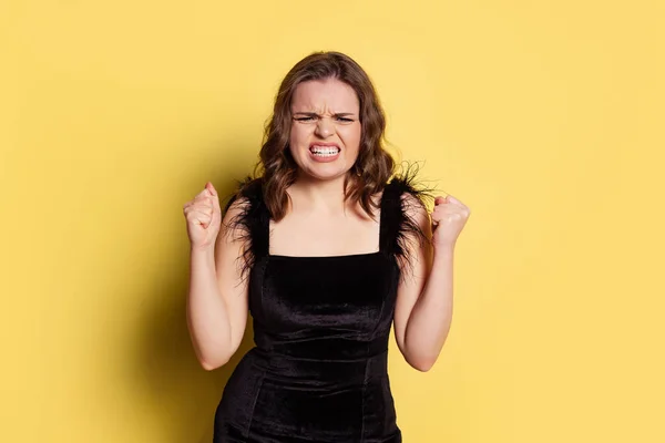 Studio colpo di giovane bella ragazza con lunghi capelli ricci che indossano abito da sera nero isolato su sfondo giallo. Concetto di emozioni, bellezza, moda — Foto Stock