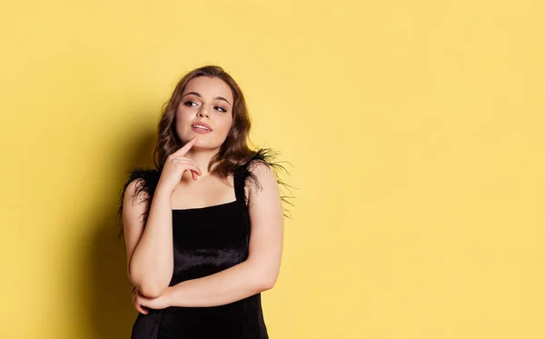 Studio shot of young pretty girl with long curly hair wearing black evening dress isolated on yellow background. Concept of emotions, beauty, fashion — Stock Photo, Image